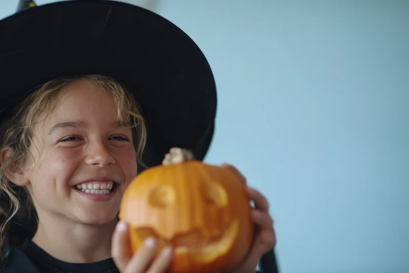 Smiling child in witch hat holding carved pumpkin for halloween celebration