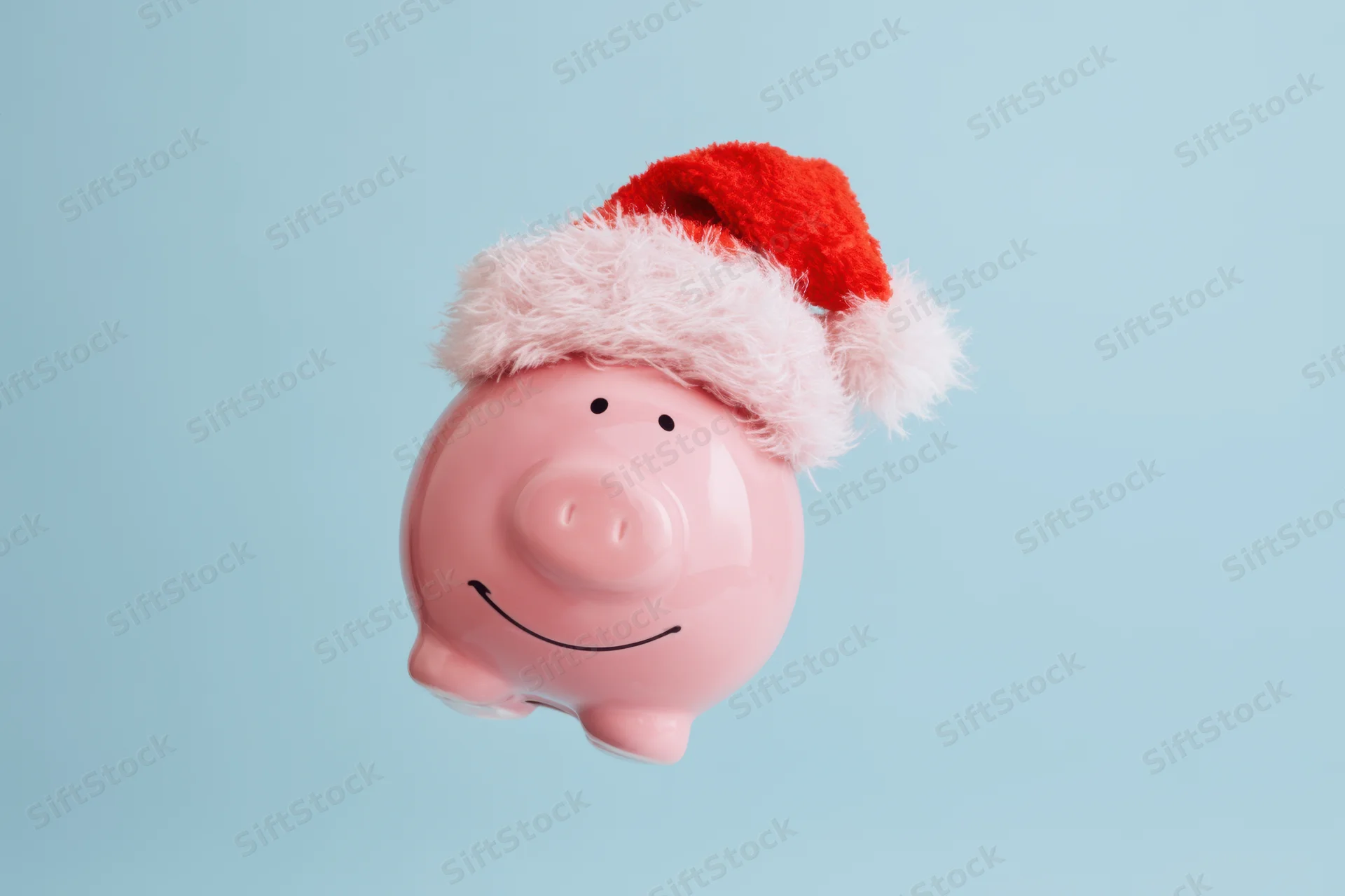 Pink piggy bank wearing santa hat smiling against light blue background