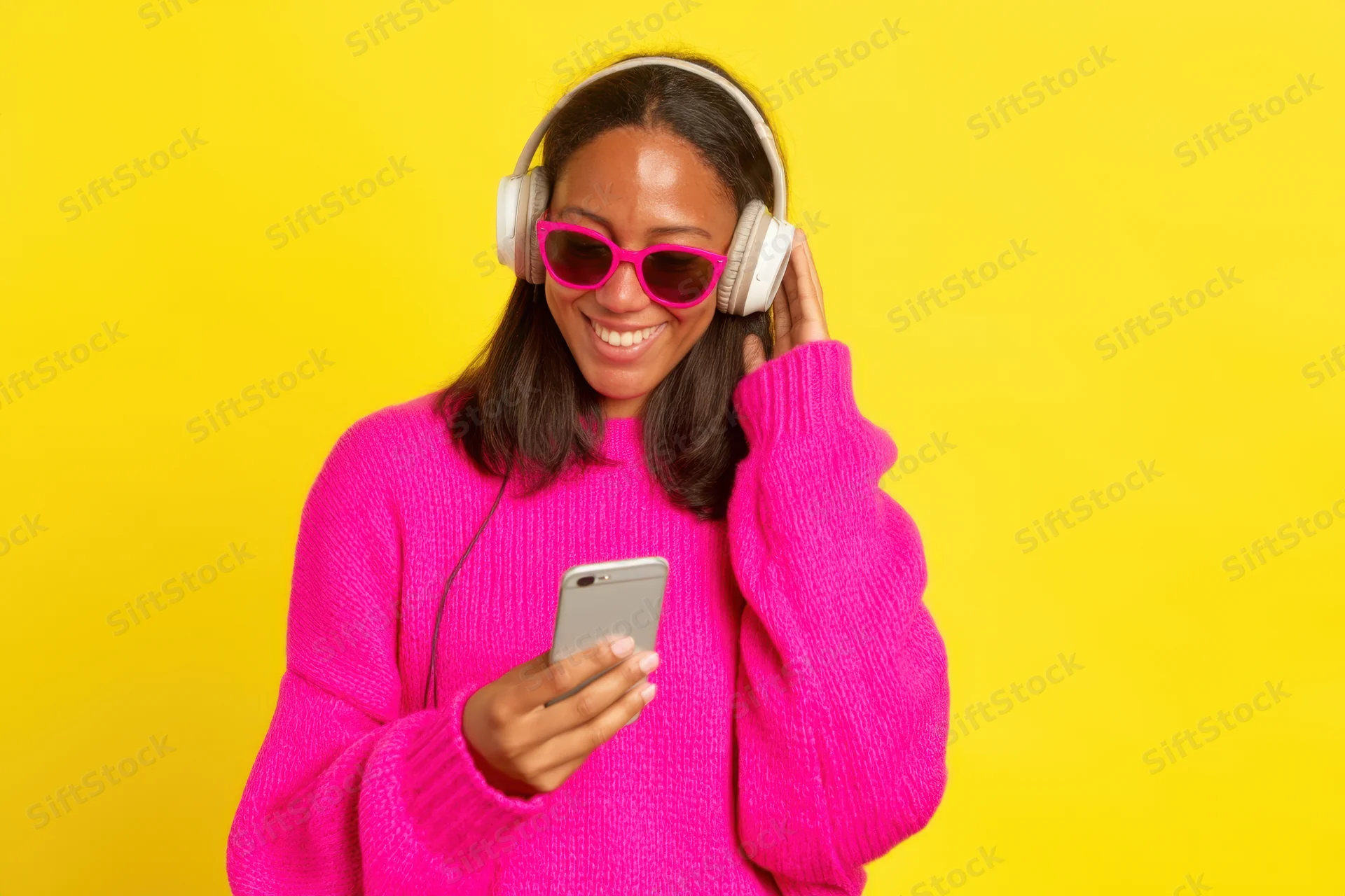 Woman smiling while listening to music on headphones and holding smartphone against yellow background