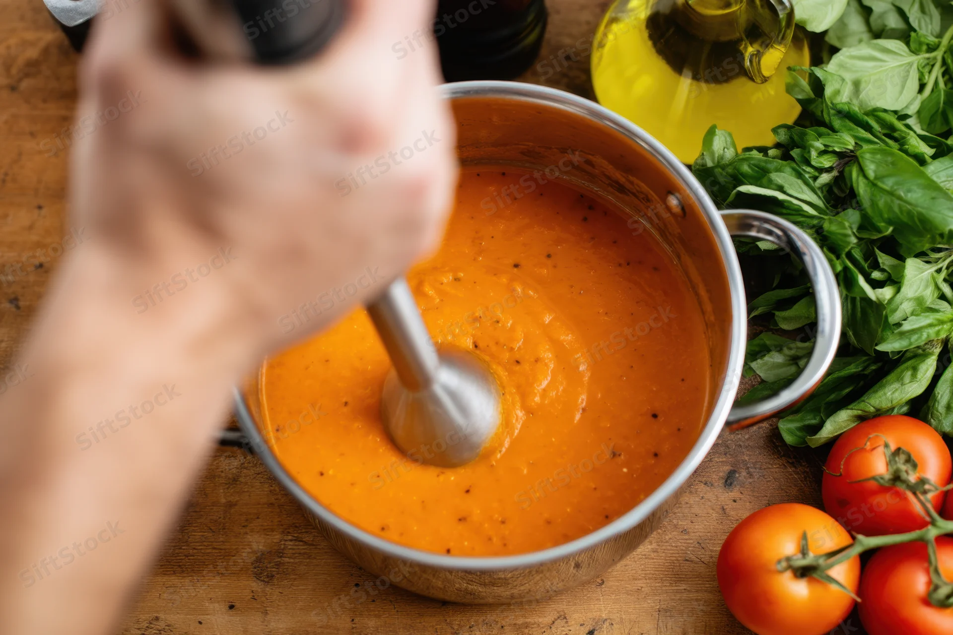 Chef using immersion blender to puree tomato sauce in a pot on a wooden countertop surrounded by fresh basil, olive oil, and tomatoes