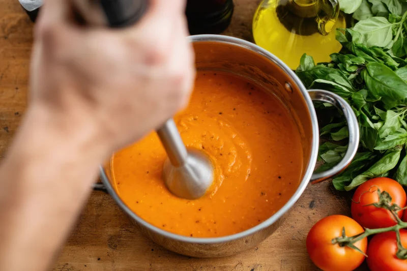 Chef using immersion blender to puree tomato sauce in a pot on a wooden countertop surrounded by fresh basil, olive oil, and tomatoes