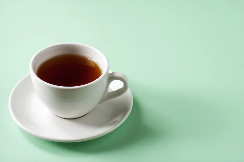 White ceramic tea cup filled with dark brown tea on saucer against mint green background