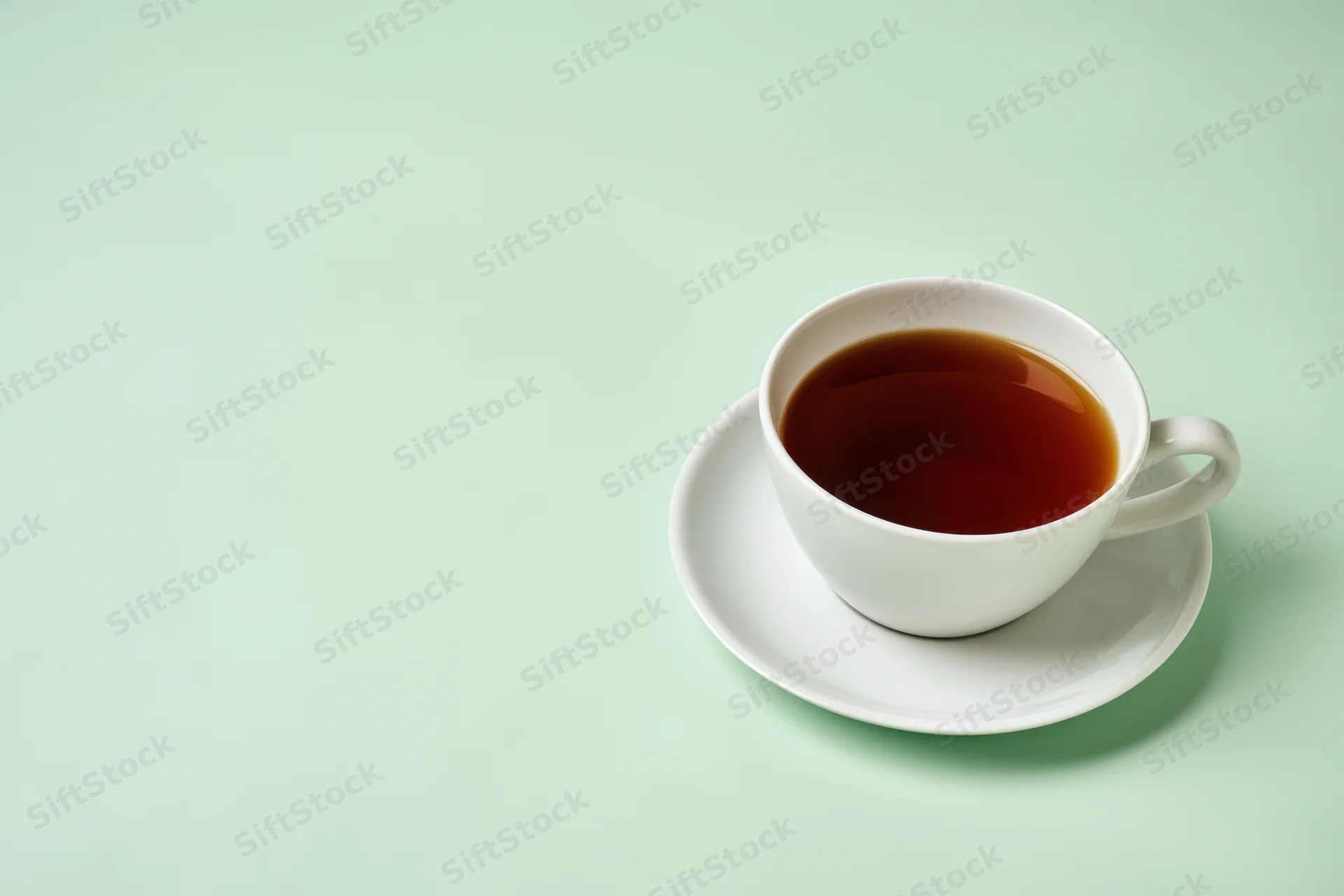 White ceramic tea cup filled with dark brown tea on saucer against mint green background