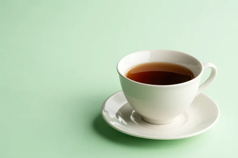 White ceramic tea cup filled with dark brown tea on saucer against mint green background
