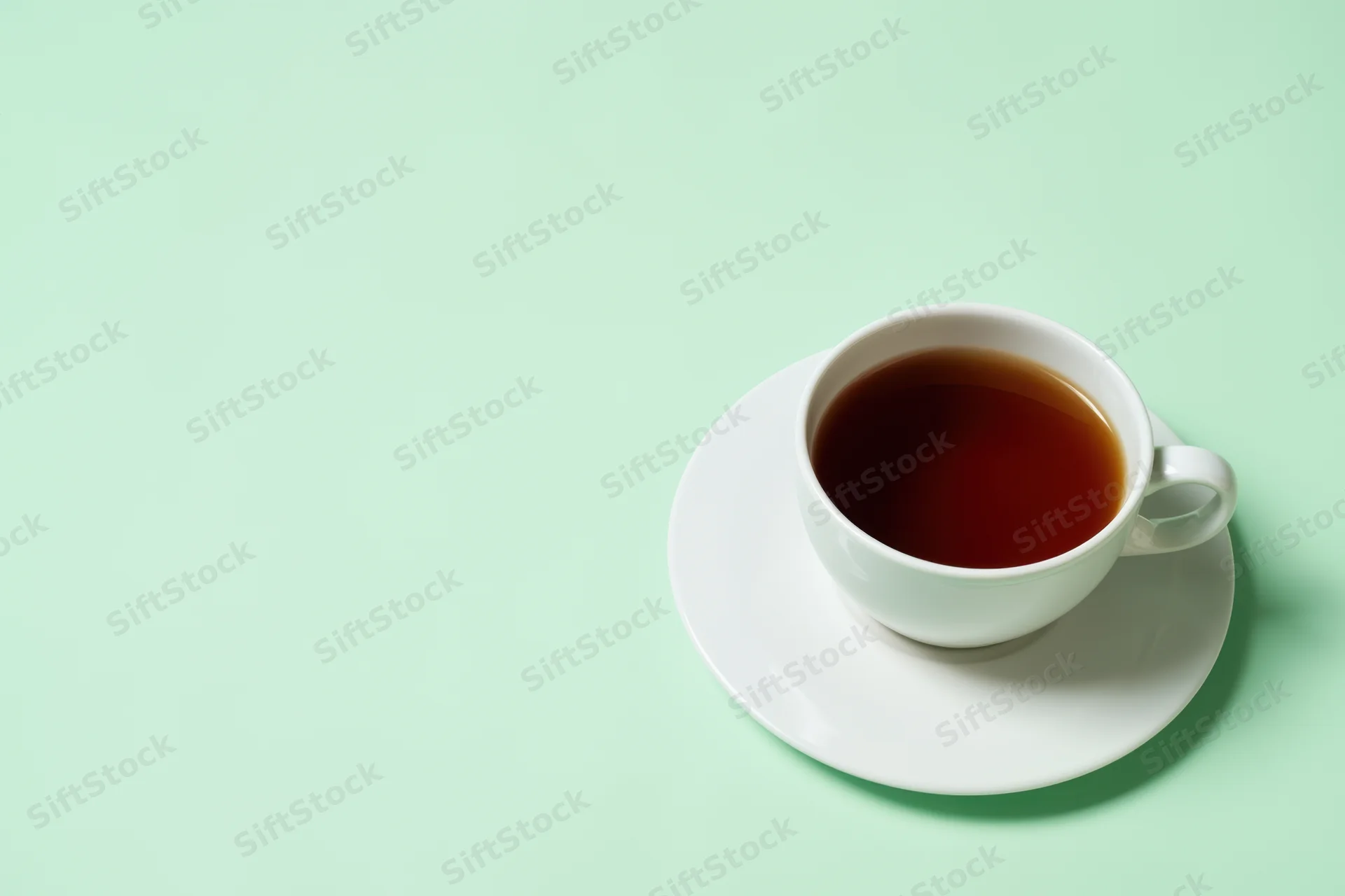 White ceramic tea cup filled with dark brown tea on saucer against mint green background with copyspace