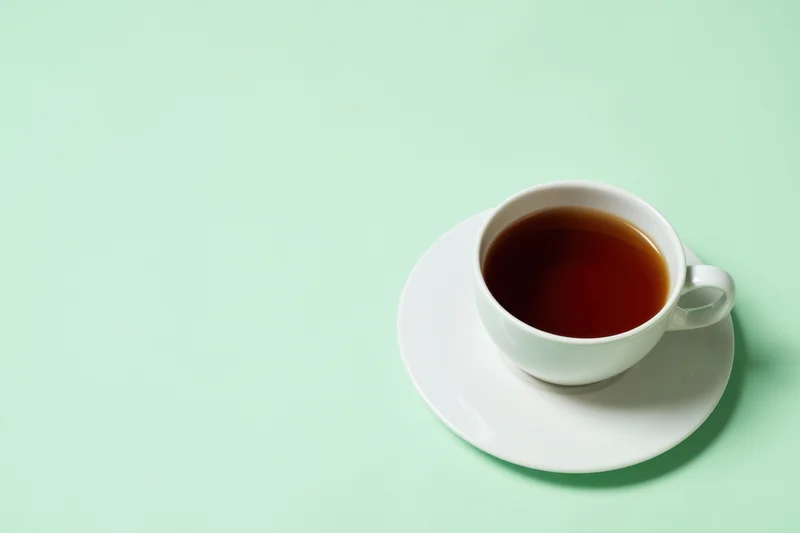 White ceramic tea cup filled with dark brown tea on saucer against mint green background with copyspace