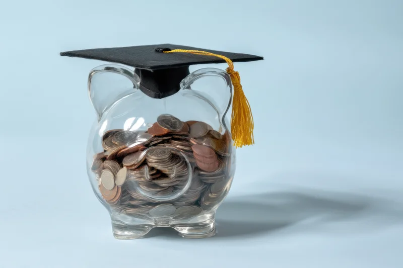 Clear glass piggy bank filled with coins topped with a graduation cap and yellow tassel on a light blue background