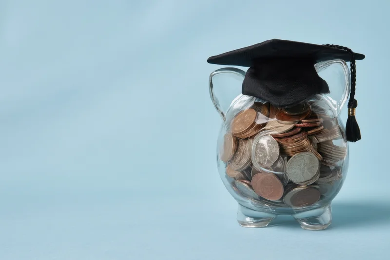 Clear piggy bank filled with coins topped with a graduation cap on a blue background