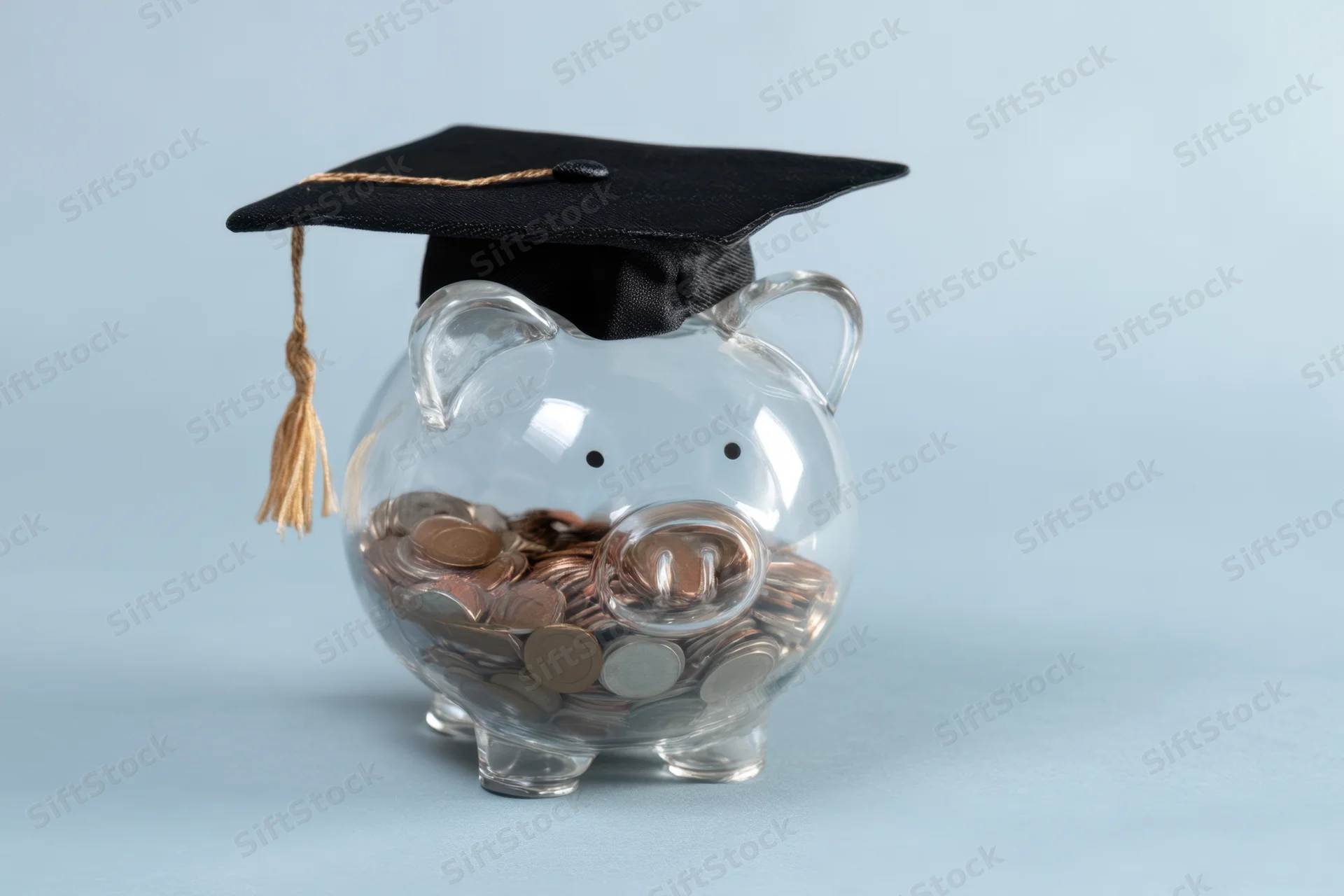 Clear glass piggy bank filled with coins wearing a black graduation cap with gold tassel on light blue background