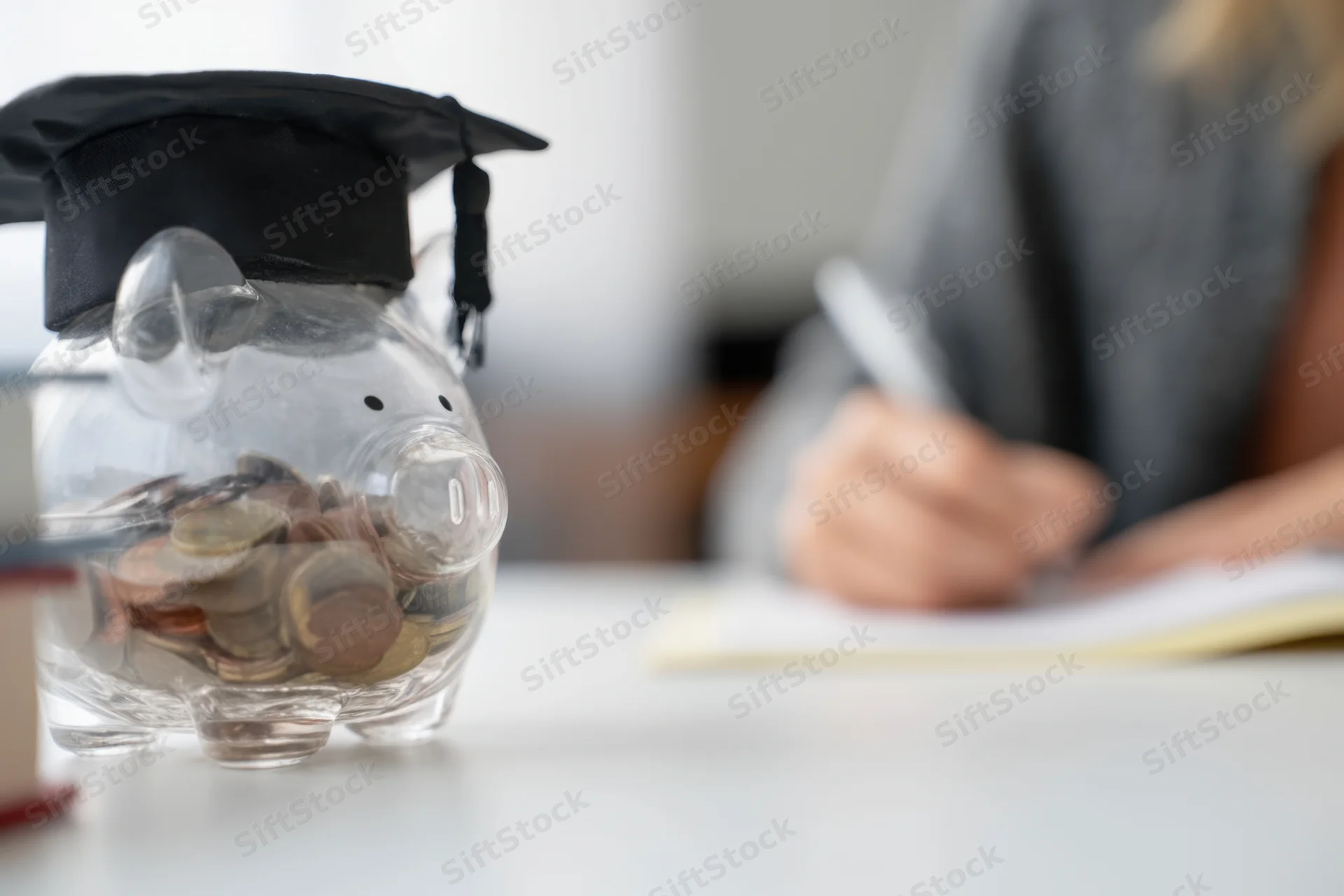 Clear piggy bank wearing graduation cap filled with coins on desk with student writing in background