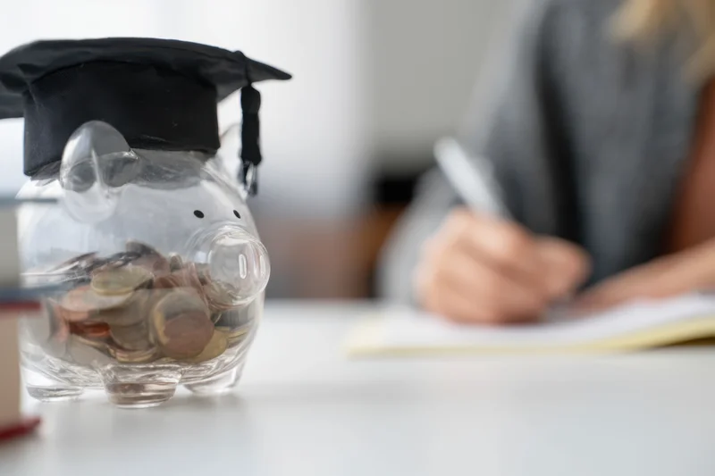 Clear piggy bank wearing graduation cap filled with coins on desk with student writing in background