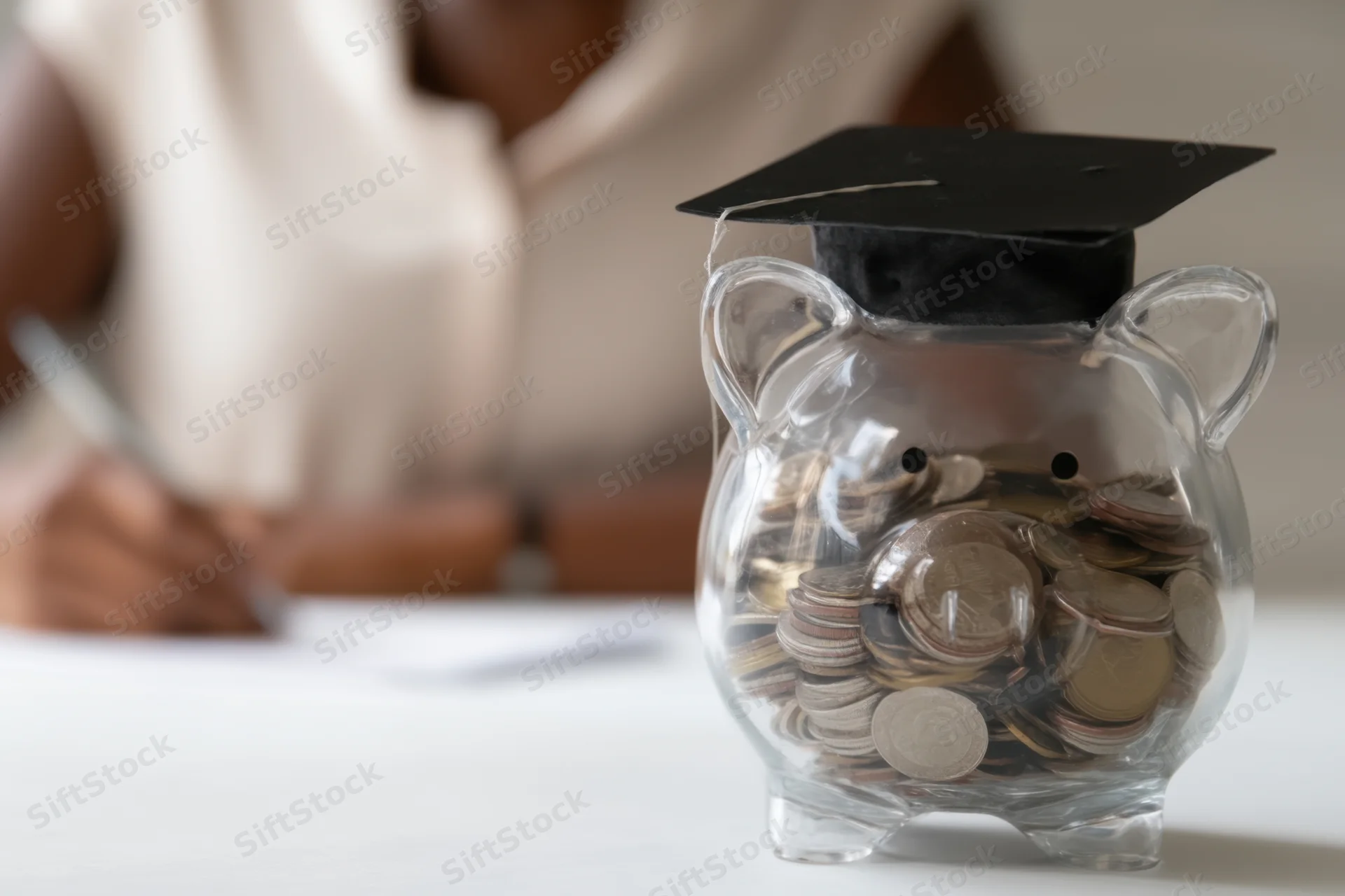 Clear piggy bank wearing graduation cap filled with coins on table with person writing in background