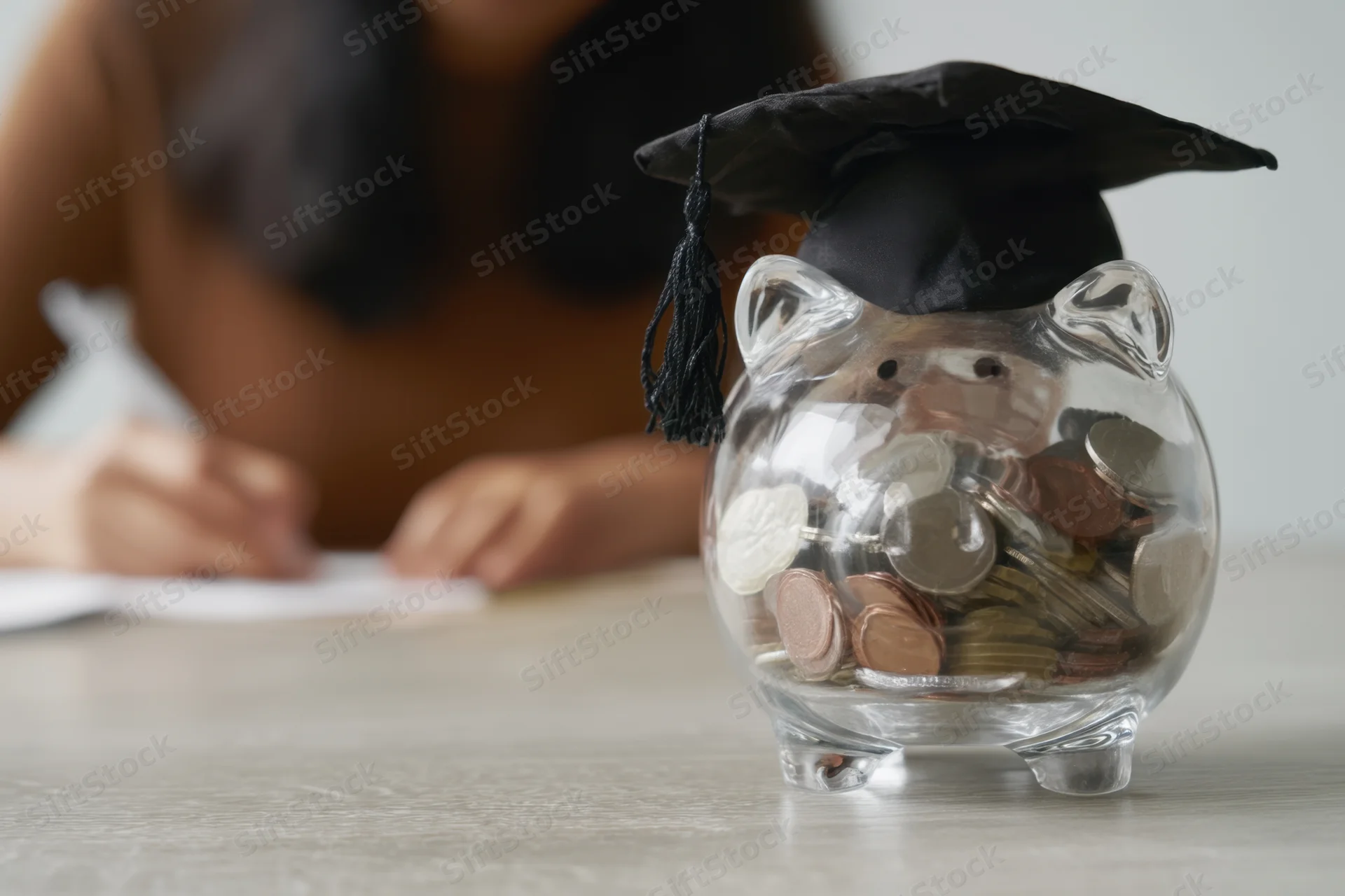 Clear glass piggy bank wearing graduation cap filled with coins on table with student writing in background