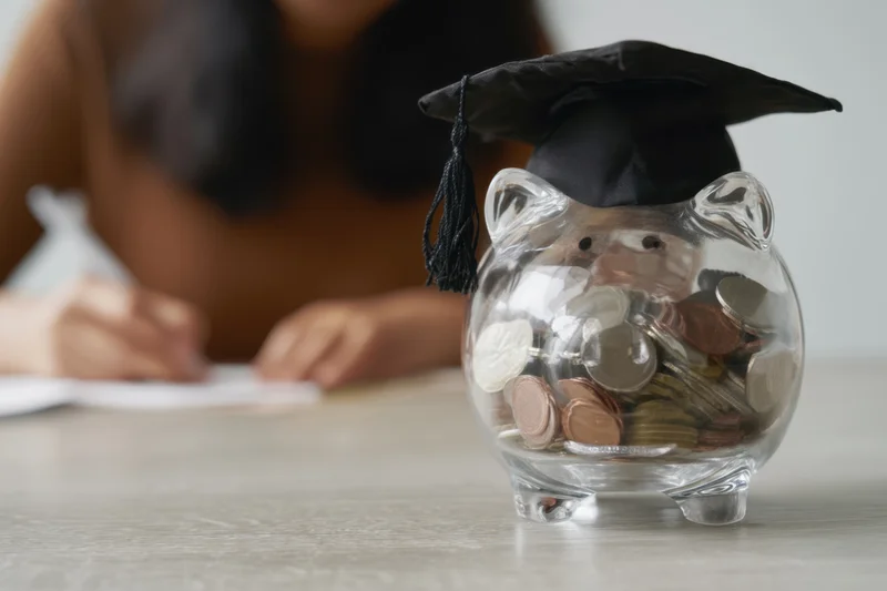 Clear glass piggy bank wearing graduation cap filled with coins on table with student writing in background