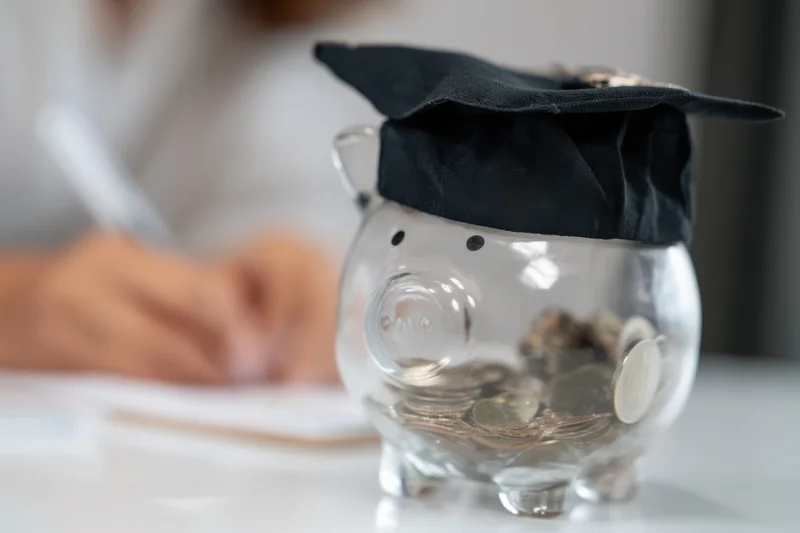 Clear glass piggy bank wearing graduation cap filled with coins on desk with person writing in background