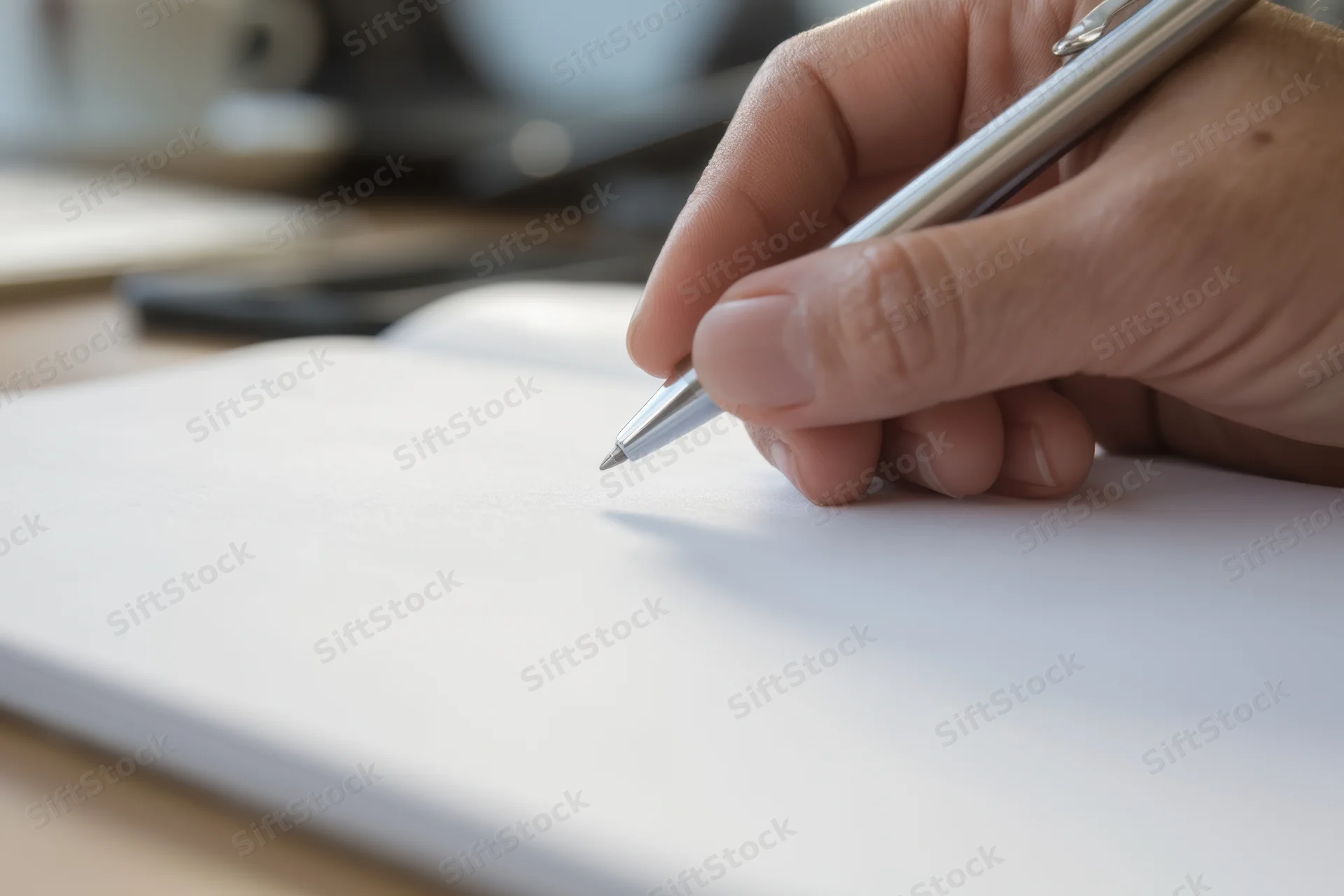Hand holding silver pen poised over blank notebook page on desk