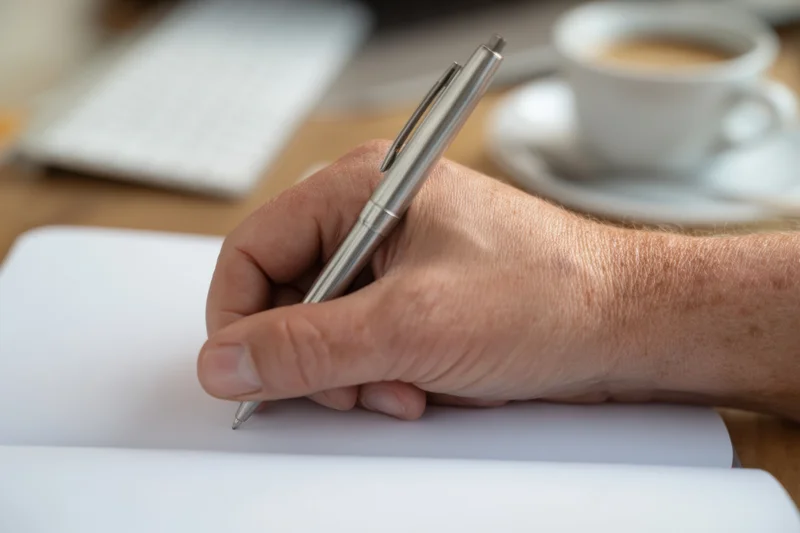 Hand holding silver pen poised over blank notebook page on wooden desk with coffee cup nearby