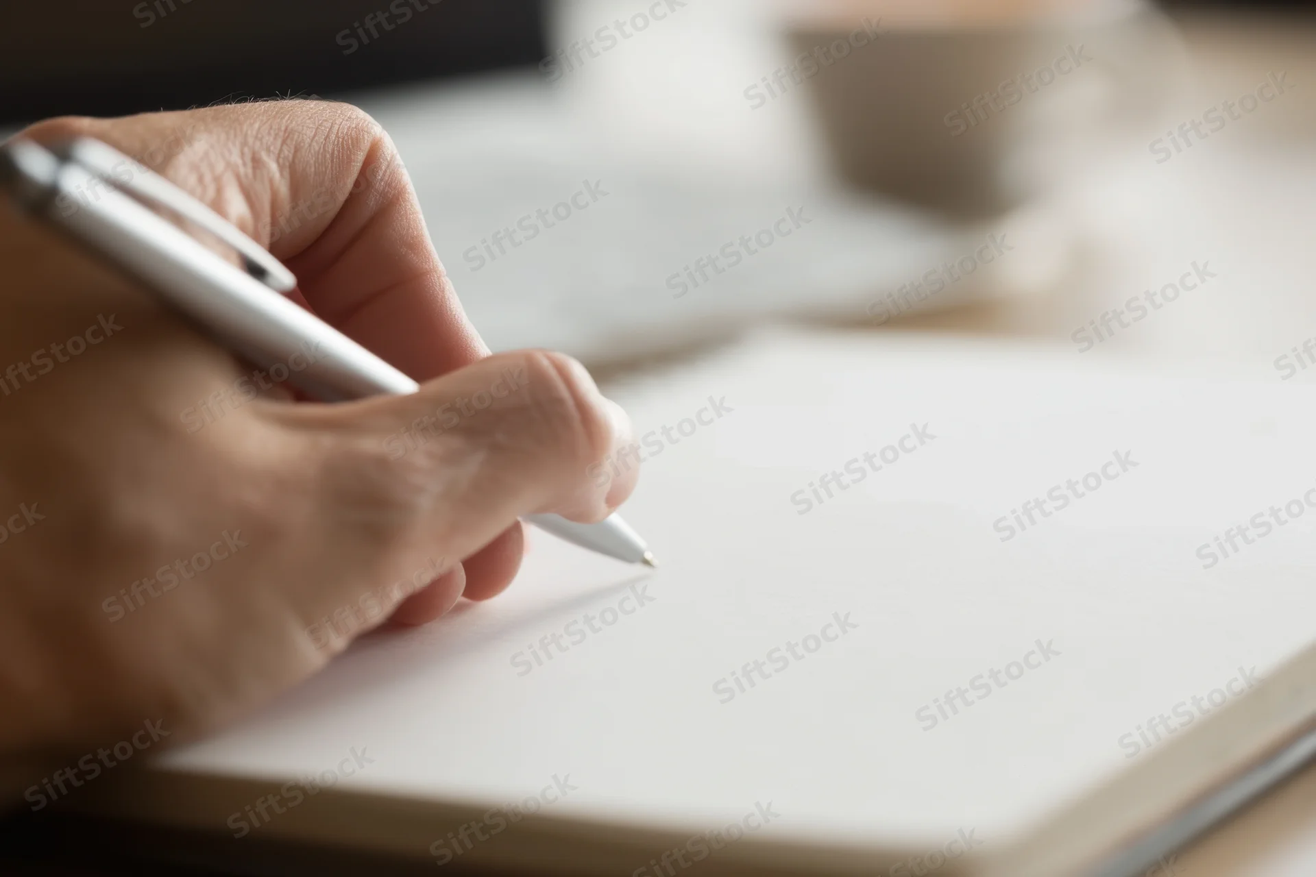 Hand holding silver pen poised over blank notebook page on desk