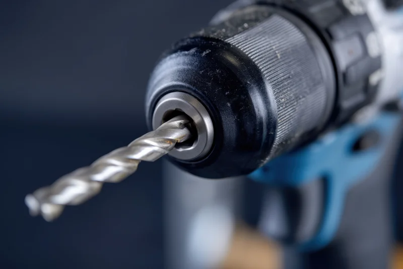 Close-up of a cordless drill with a metallic drill bit inserted in the chuck, showing the grip and battery housing against a blurred background
