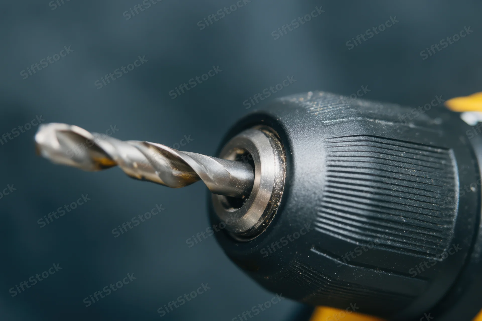 Close-up of a cordless drill with a metallic drill bit inserted in the chuck, showing textured grip and yellow accents