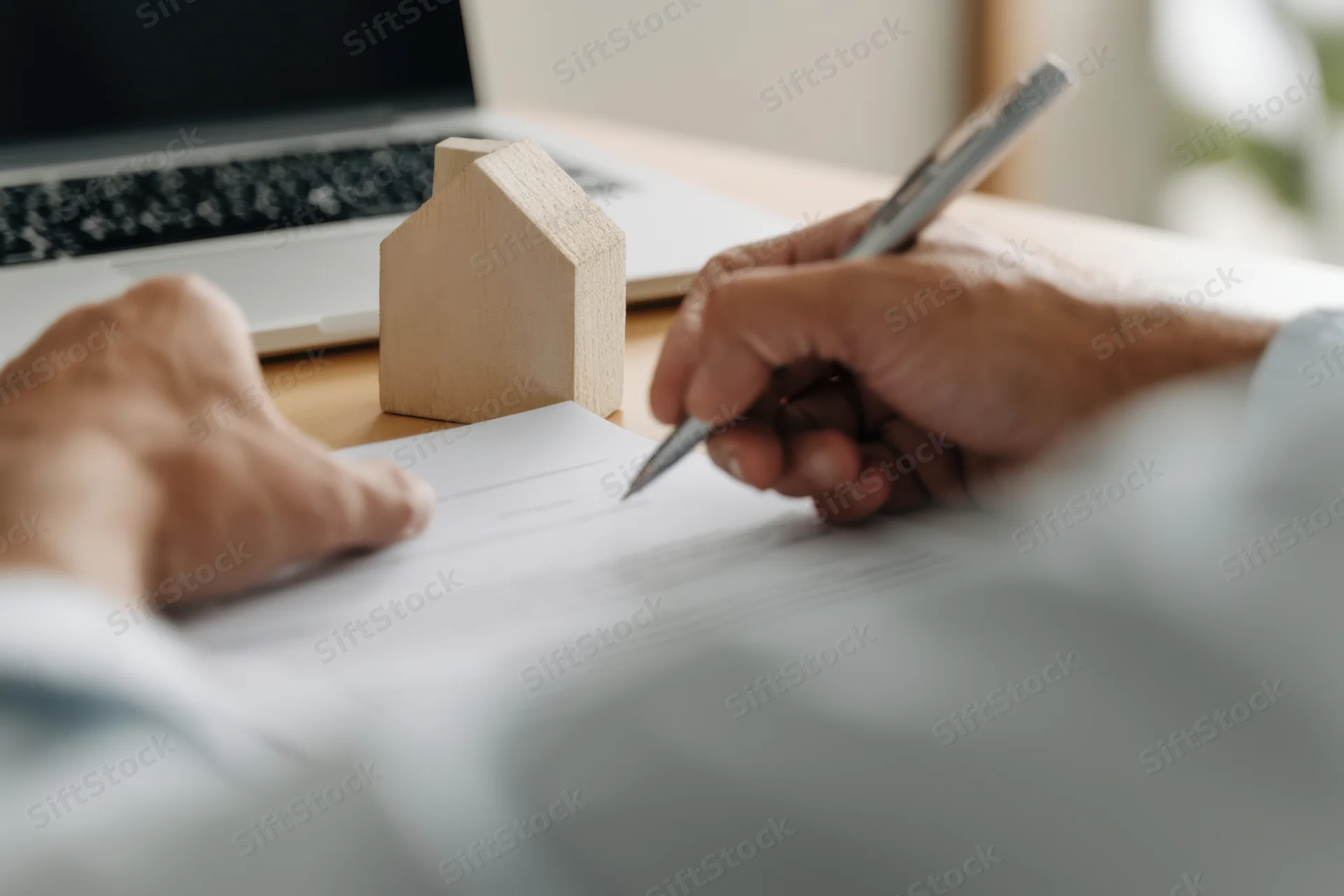 Hands signing a document on a desk with a wooden house model and laptop nearby