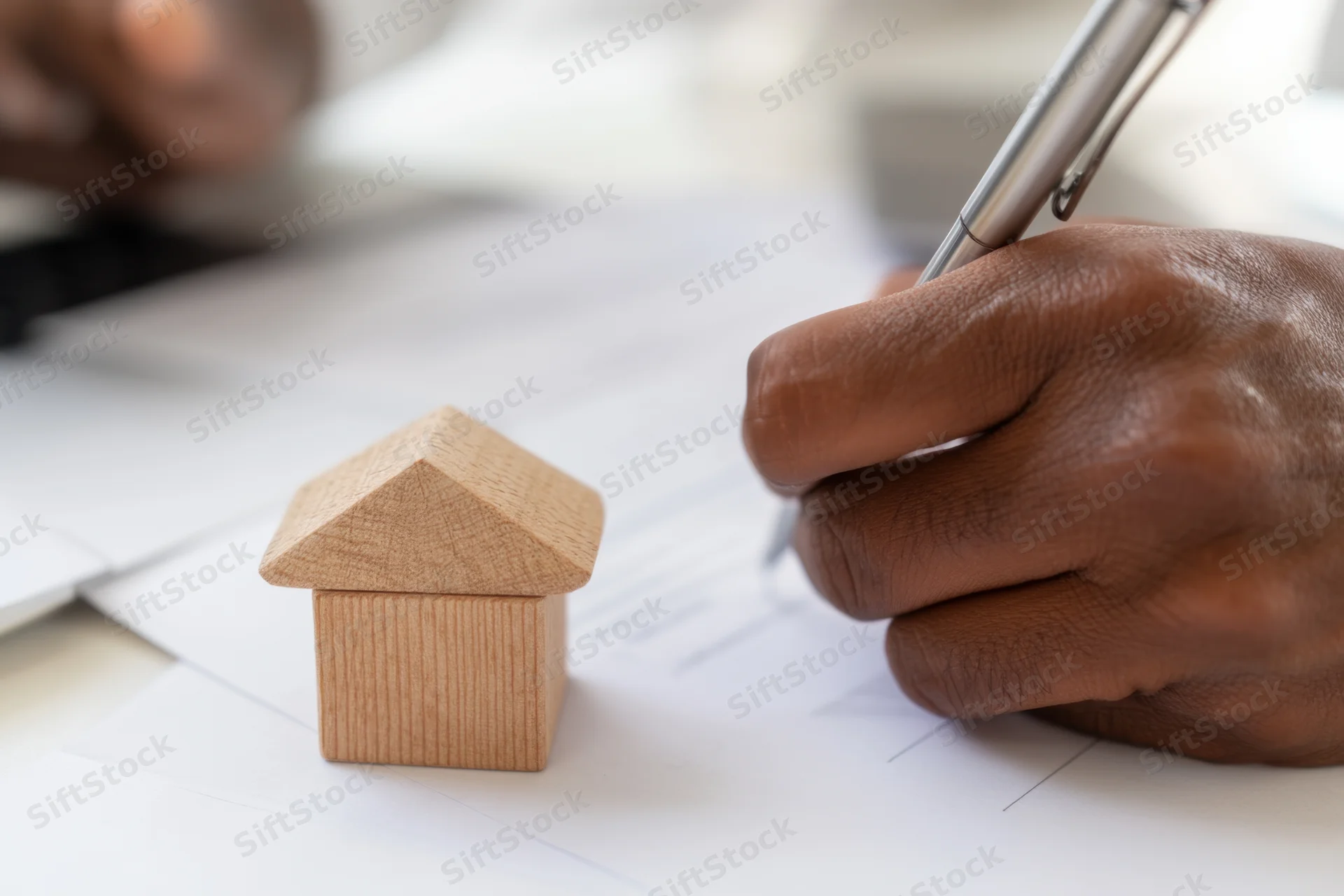 Hand signing a document next to a wooden house model on a desk