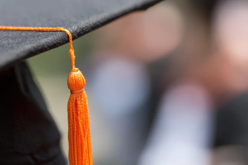 Close-up of a black graduation cap with an orange tassel hanging from the edge, blurred background showing indistinct figures