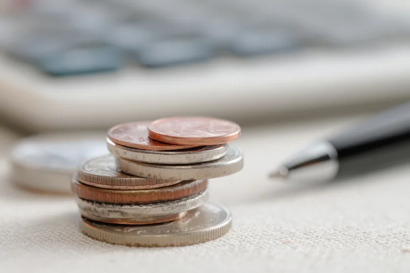 Stack of coins with various denominations on a desk near a calculator and pen