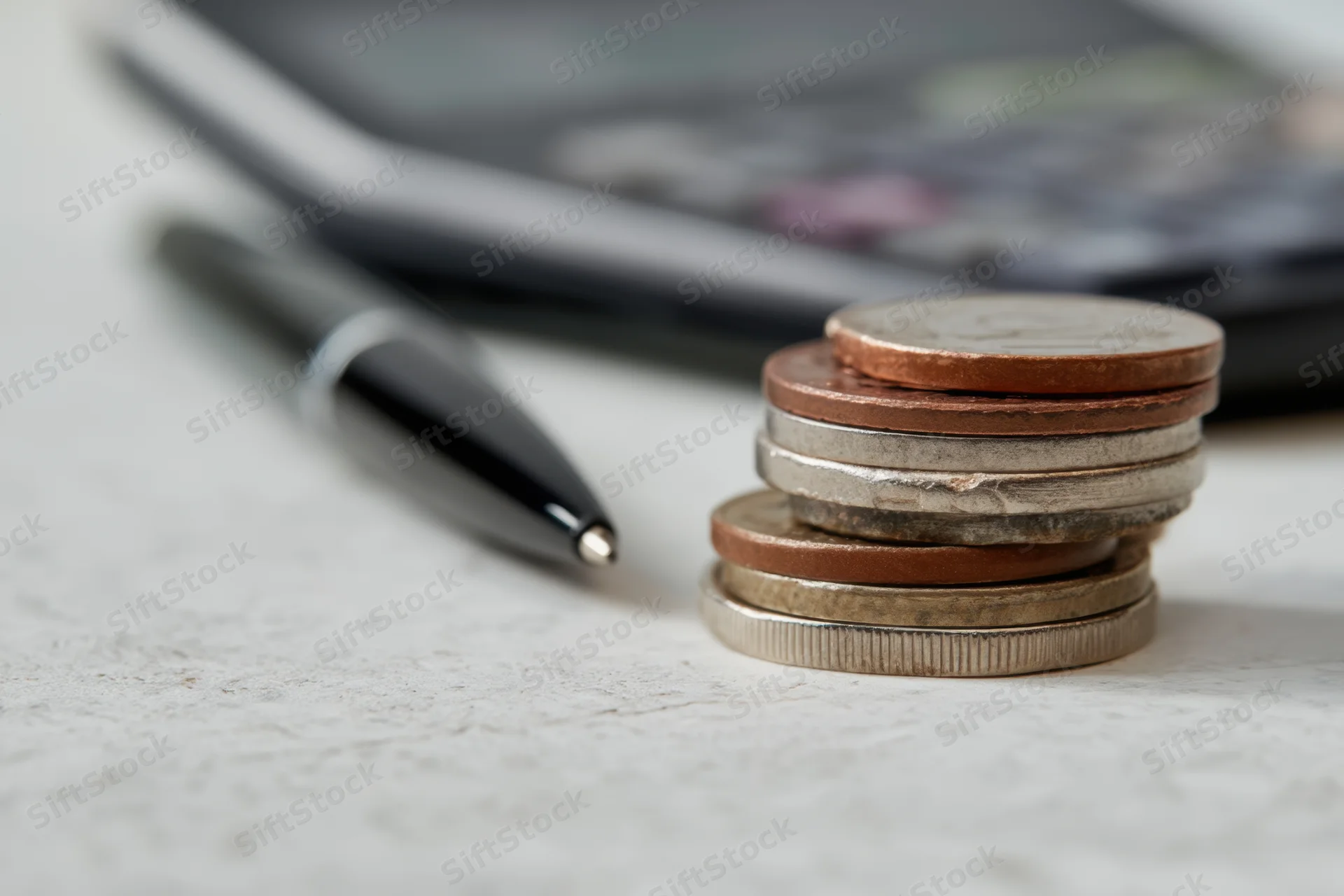Stack of coins with various denominations on a desk near a calculator and pen