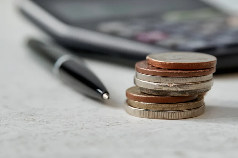 Stack of coins with various denominations on a desk near a calculator and pen
