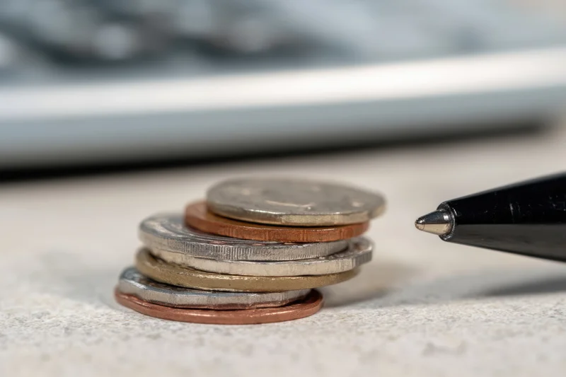 Stack of coins with various denominations on a desk surface next to a pen