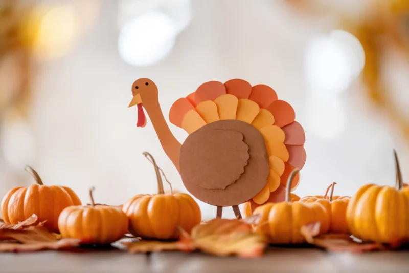 Paper turkey standing on a table surrounded by small pumpkins and autumn leaves
