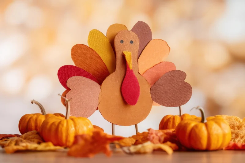 Paper turkey craft standing on table surrounded by pumpkins and autumn leaves