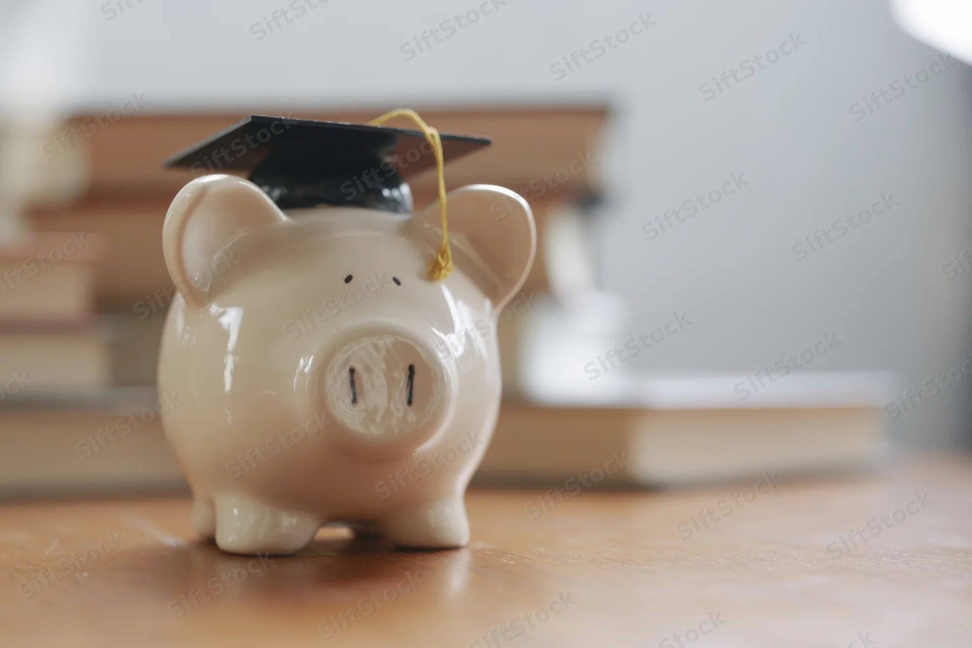 Ceramic piggy bank wearing graduation cap on wooden desk with books in background