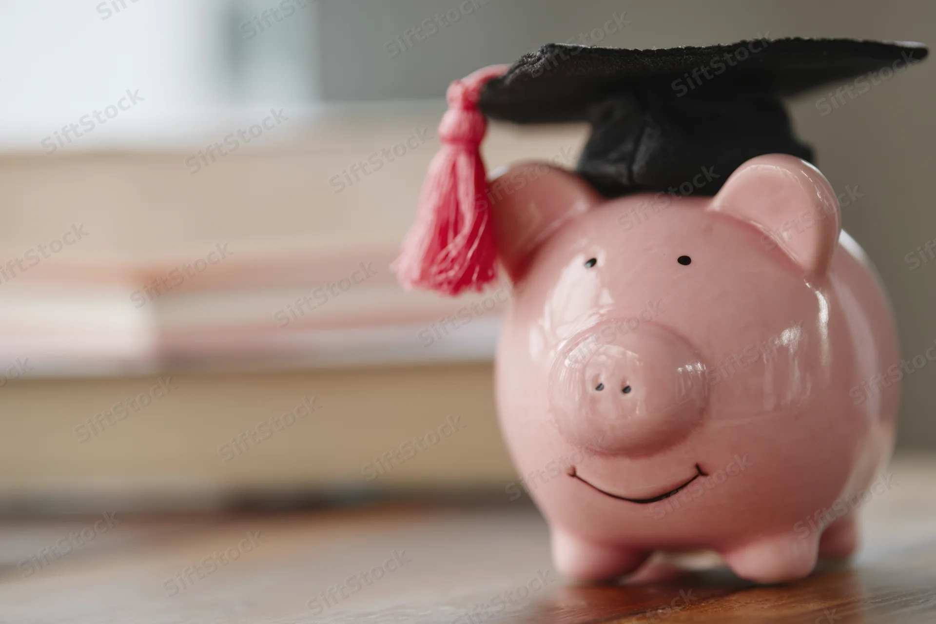 Pink ceramic piggy bank wearing black graduation cap with pink tassel on wooden surface near stack of books