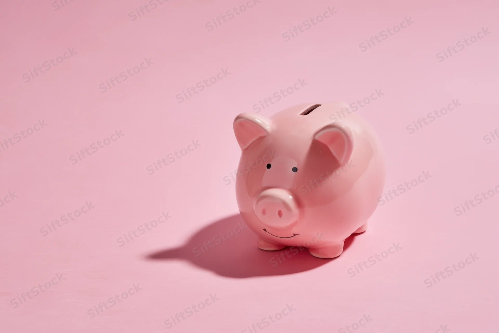 Pink ceramic piggy bank with coin slot on soft pink background casting shadow