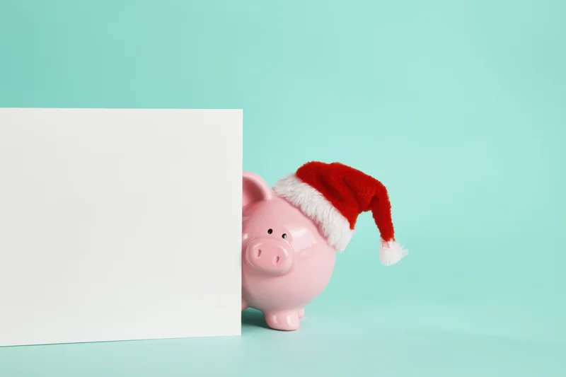 Pink piggy bank wearing santa hat peeking from behind white card on teal background