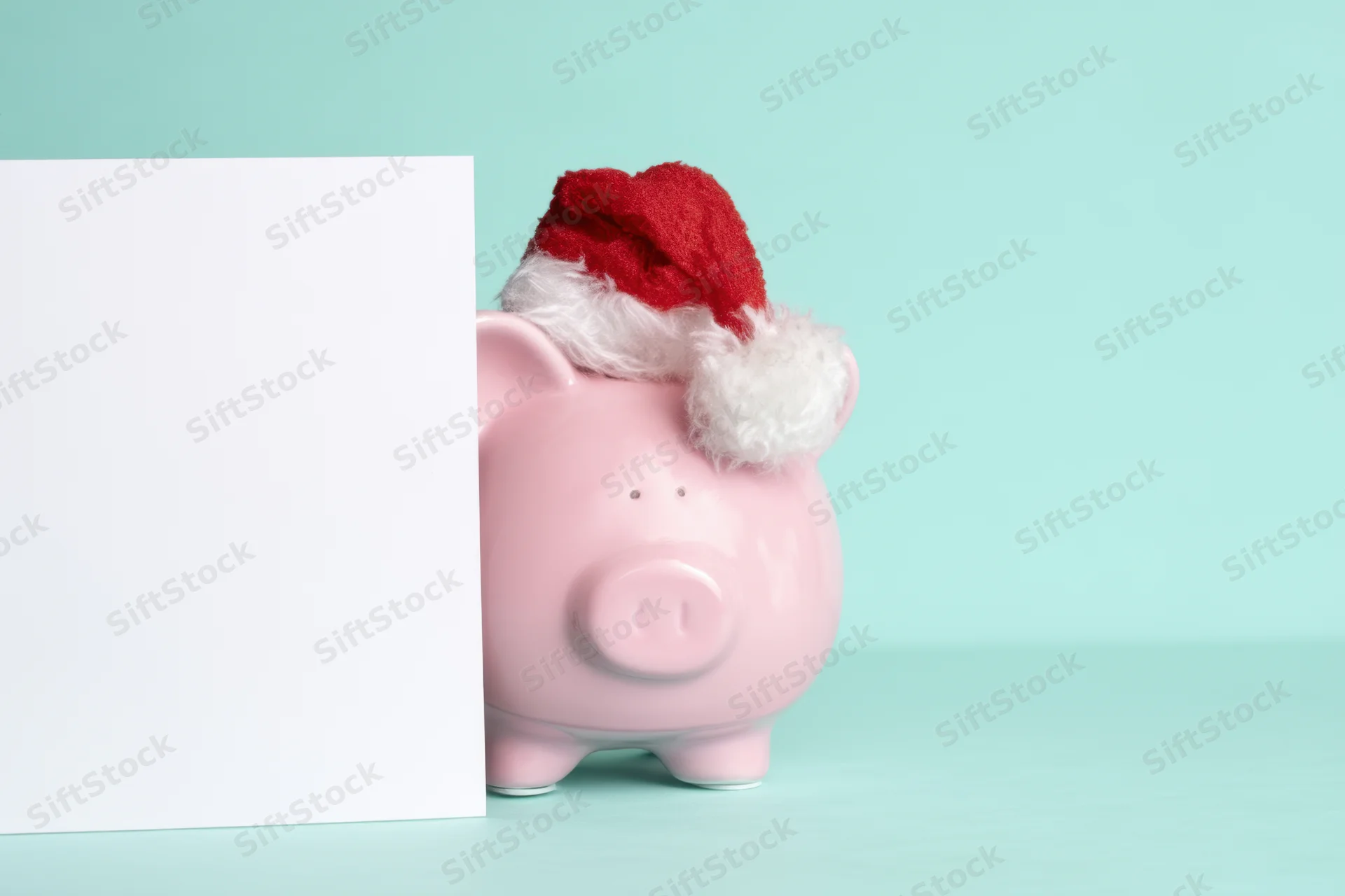 Pink piggy bank wearing santa hat peeking from behind white card on mint background