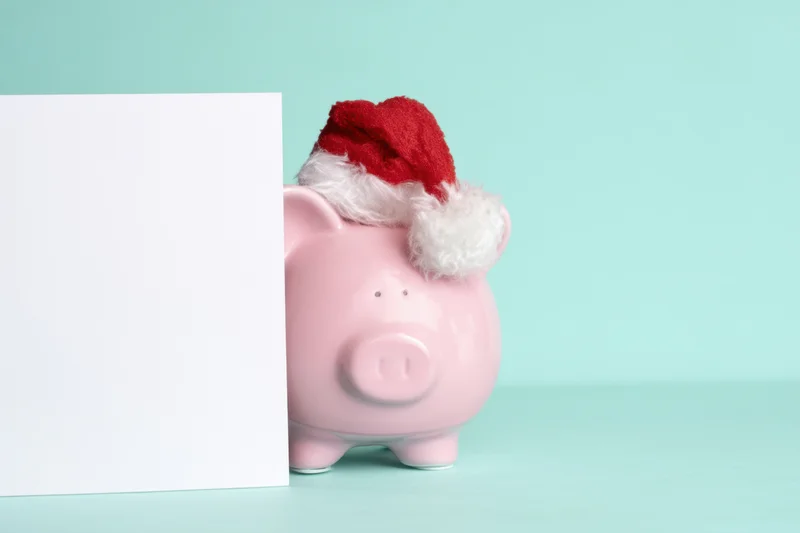 Pink piggy bank wearing santa hat peeking from behind white card on mint background