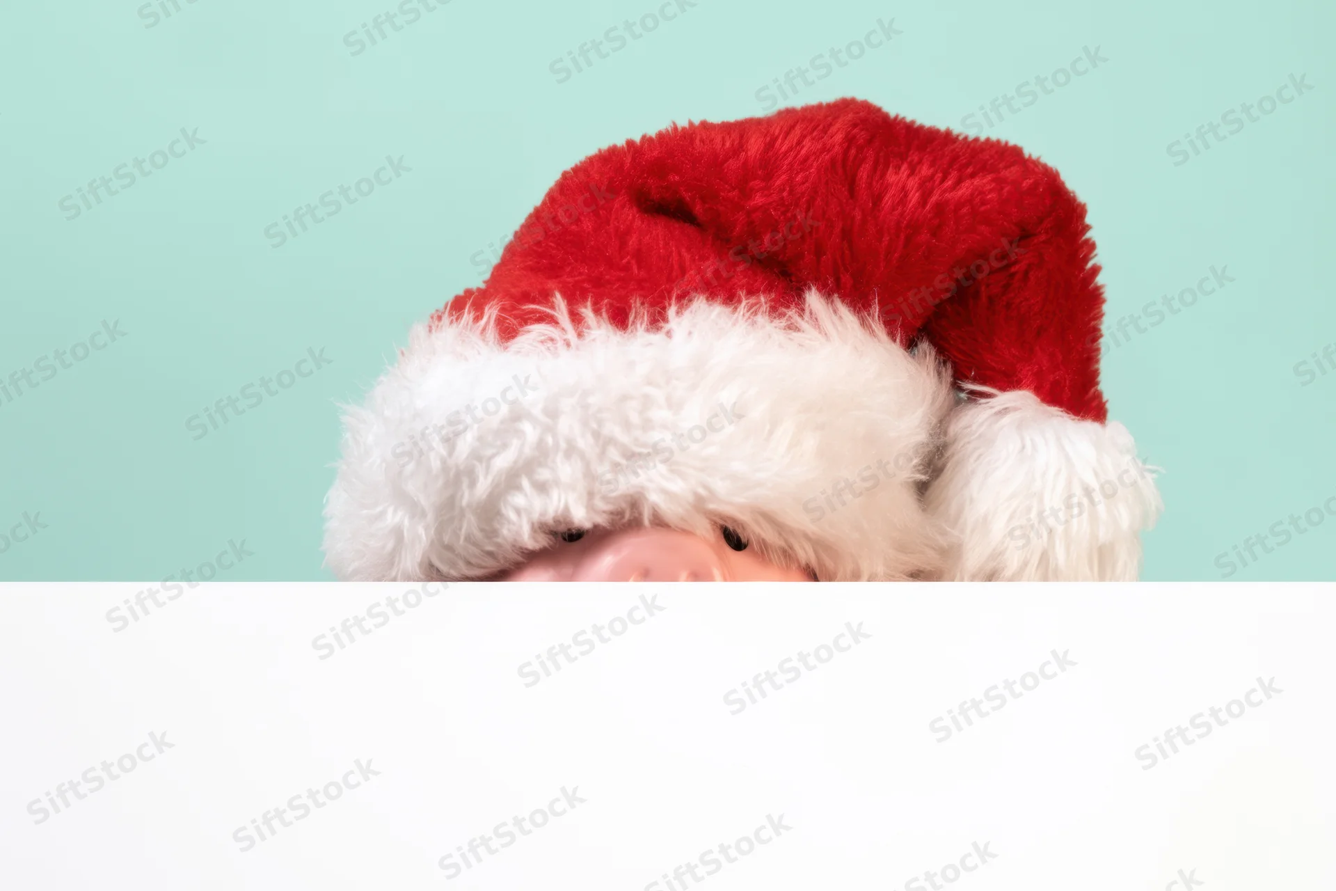 Pink piggy bank wearing santa hat peeking over white banner against teal background