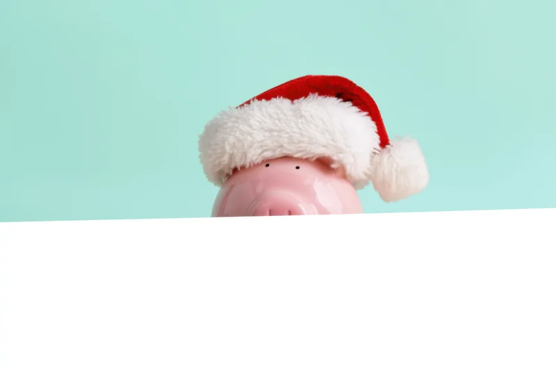 Pink piggy bank wearing santa hat peeking over white banner against teal background