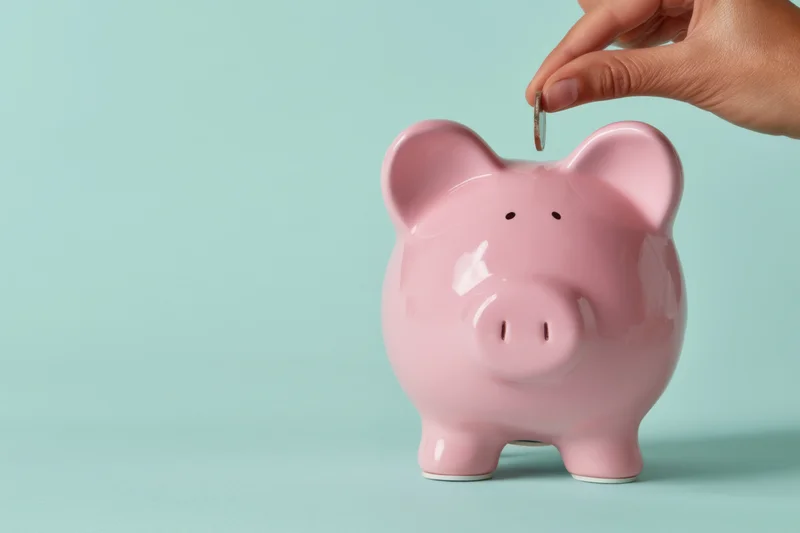 Hand dropping a coin into a pink piggy bank on a light blue background