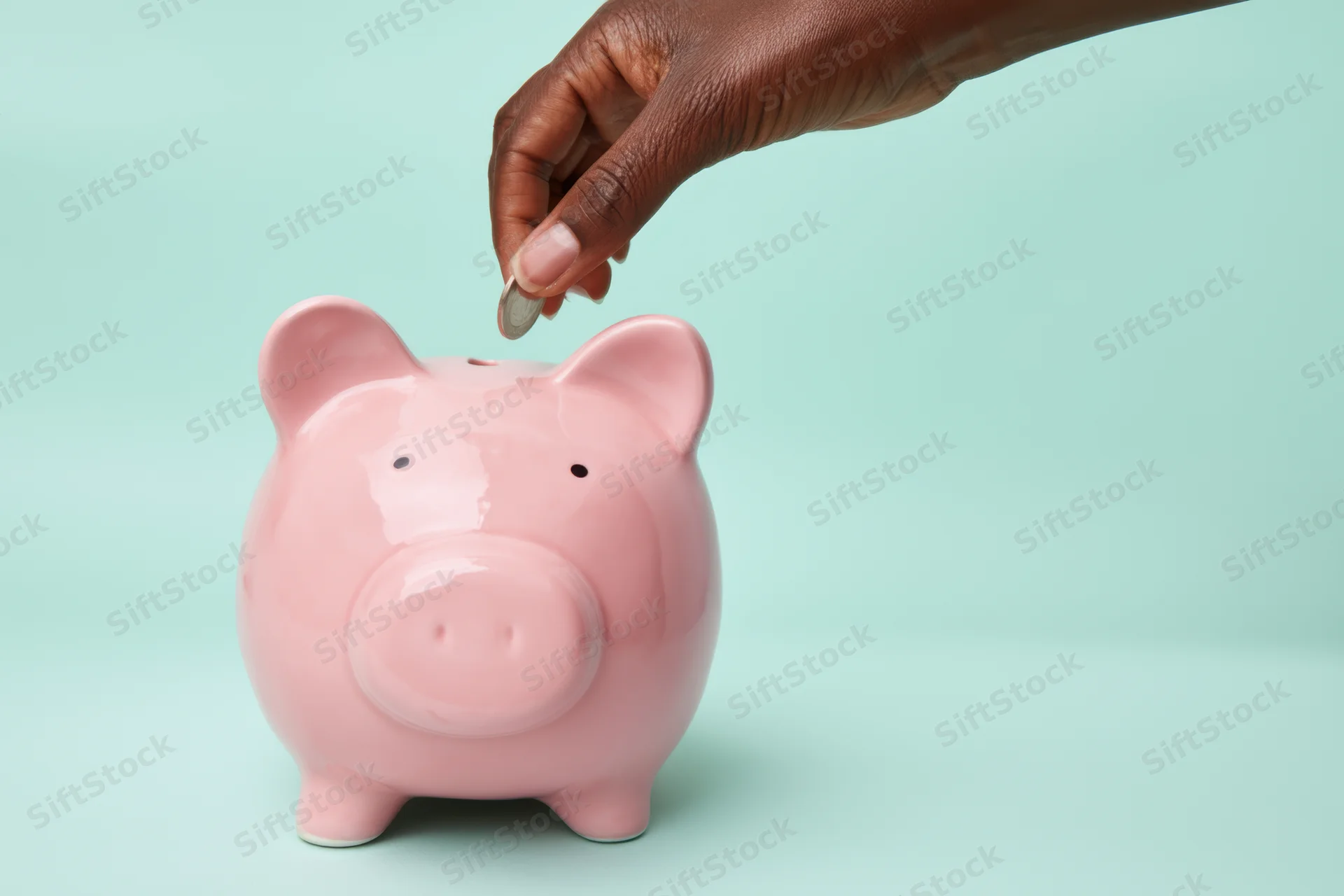 Hand dropping a coin into a pink ceramic piggy bank on a mint background