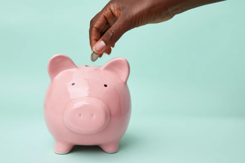 Hand dropping a coin into a pink ceramic piggy bank on a mint background