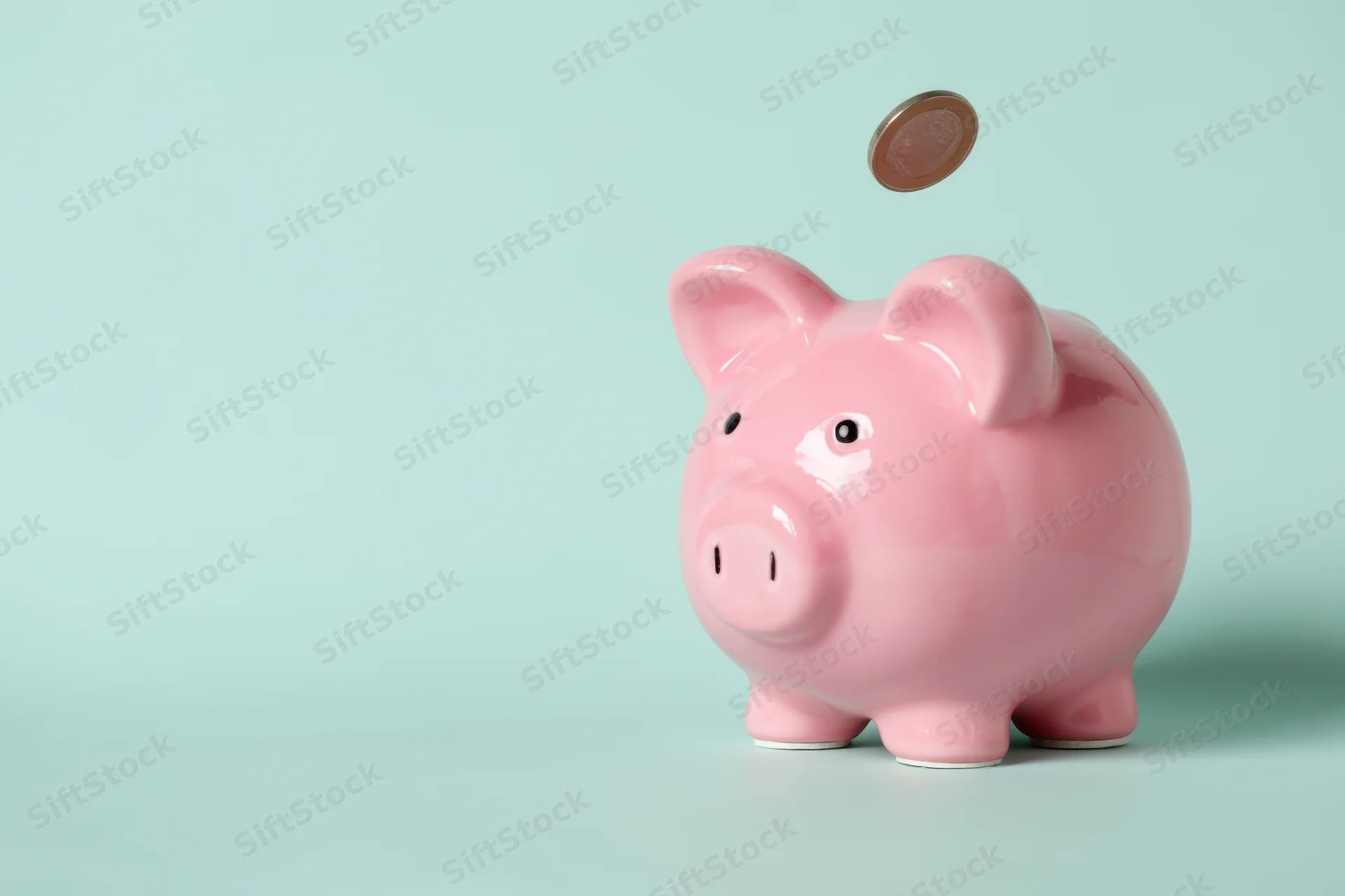 Pink ceramic piggy bank with coin dropping into slot on light blue background