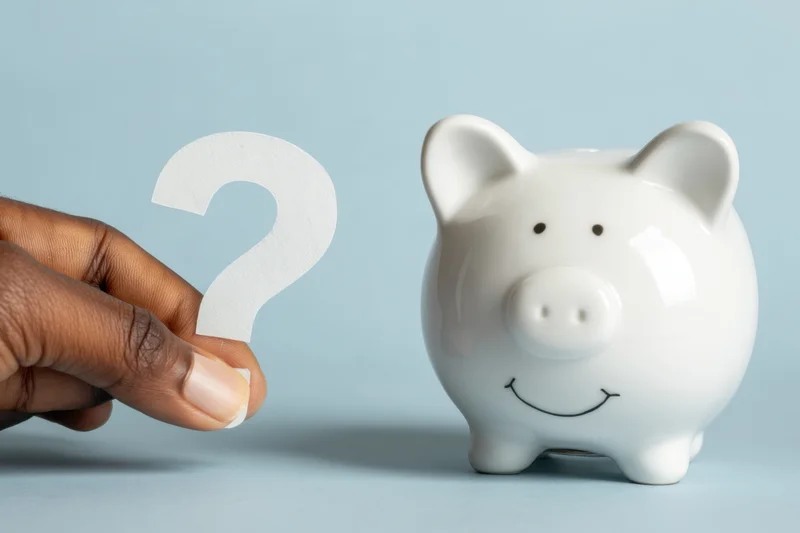 White ceramic piggy bank with smiling face beside a hand holding a paper question mark on a blue surface