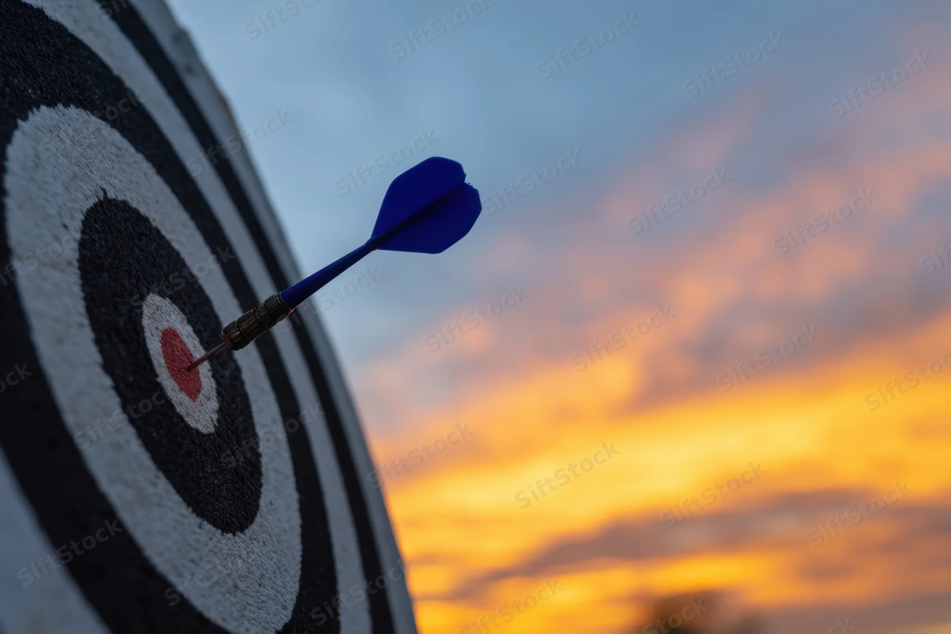 Blue dart striking the bullseye center of a dartboard against a sunset sky