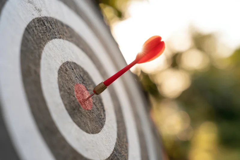 Red dart striking the center of a dartboard outdoors in sunlight