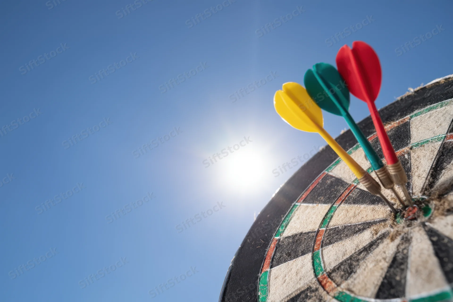 Three colorful darts hitting the bullseye of a dartboard against a bright blue sky with sun