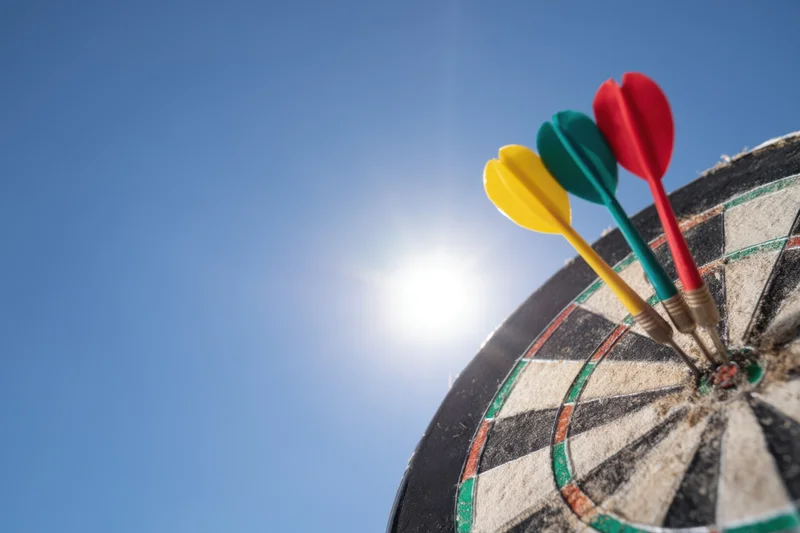 Three colorful darts hitting the bullseye of a dartboard against a bright blue sky with sun